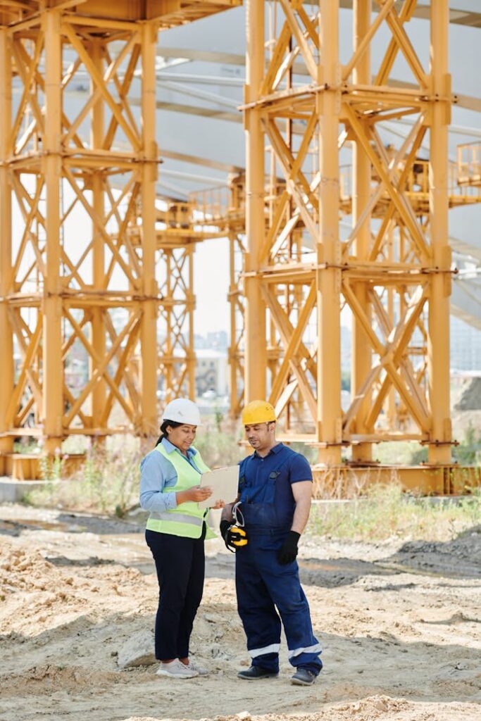 Two construction workers examining plans at a site with large steel structures.